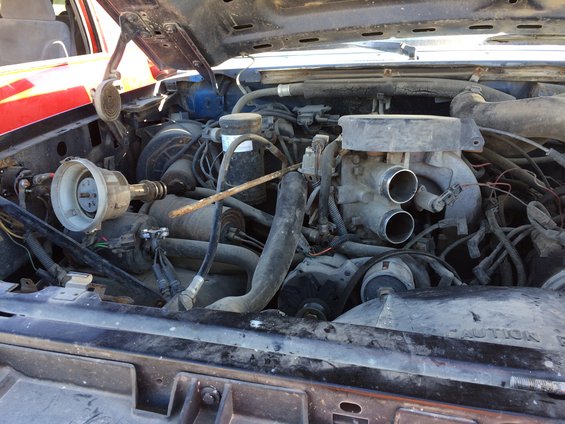 Engine bay with distributor cap removed, showing carburetor and hoses. Red vehicle fender visible on left