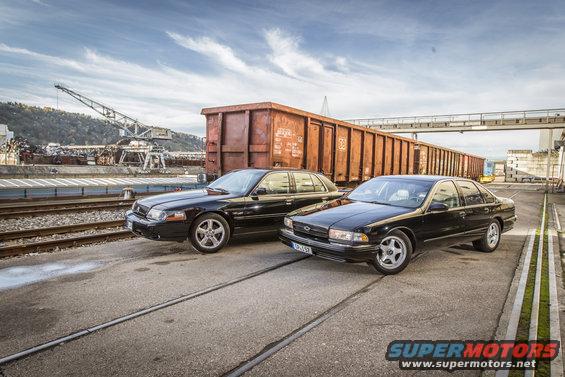Two black sedans, one a 1996 Chevrolet Impala SS, parked side-by-side on train tracks near a freight train car