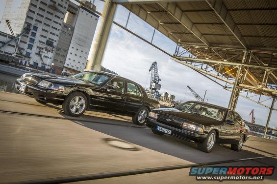 Two black 2003 Mercury Marauders driving side-by-side under an overpass with industrial buildings and cranes in the background