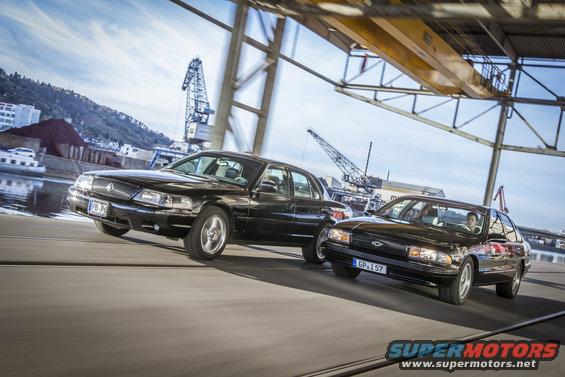 Two black sedans driving on a road near a harbor with cranes