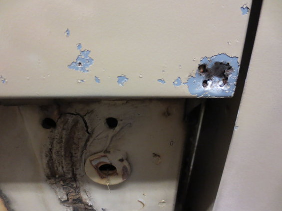 Close-up of a beige vehicle panel with chipped paint revealing blue primer and rust spots, showing some undercarriage detail