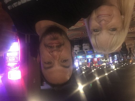 Two people pose for a selfie in front of illuminated slot machines inside a casino