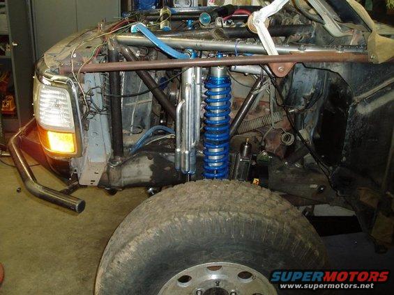 Off-road race truck front end with custom roll cage, blue coilover shocks, and large tire. Headlight visible