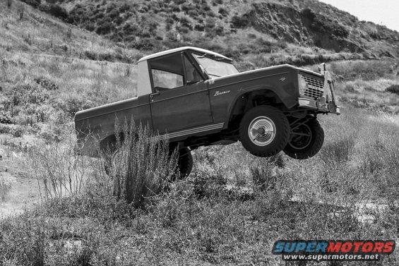 Ford Bronco airborne over grassy terrain. The vehicle is dark colored with a white top and has the word "Bronco" on the side