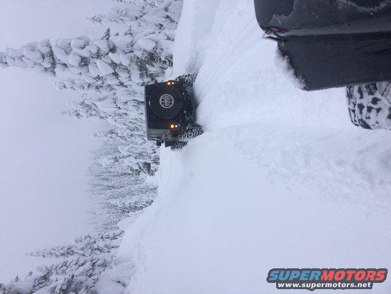 Black Jeep Wrangler driving on a snowy mountain road, with another vehicle partially visible on the right