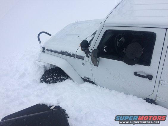 White Jeep Wrangler stuck in deep snow, front fender and tire visible, mountain decal on hood