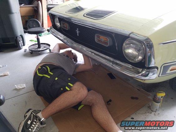 Man lying on cardboard working on the front underside of a 1971 Ford Mustang