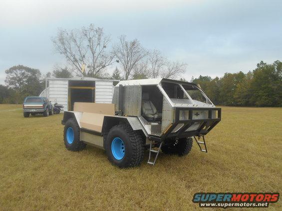 Custom off-road vehicle with large blue wheels and a metal body, parked in a grassy field next to a building. Another SUV is visible in the background