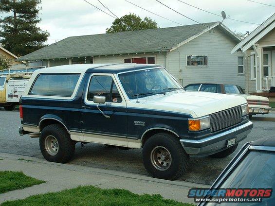 Two-tone blue and white 1990 Ford Bronco parked on street, other vehicles visible
