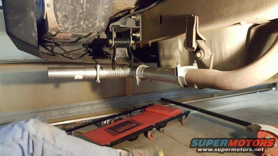 Underside of a vehicle showing exhaust pipe repair with a new flex pipe and clamps. A red creeper is on the floor