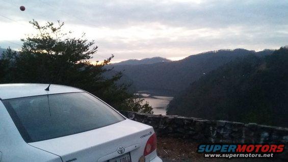 White sedan parked on a scenic overlook with a lake and mountains in the background