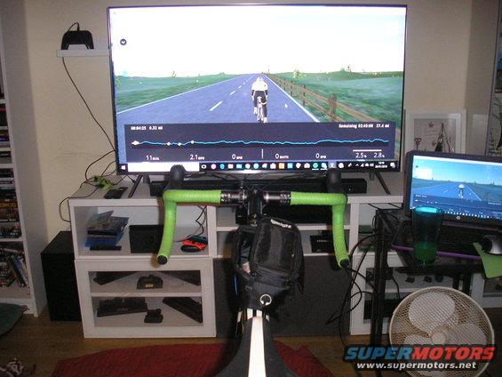 Indoor bicycle trainer with handlebars, large monitor displaying a cycling simulation, and a laptop
