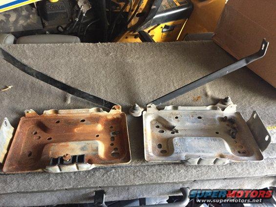 Two metal battery trays, one rusted, with black hold-down brackets. Parts are on a carpeted surface
