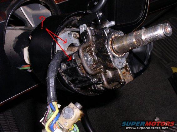 Close-up of a vehicle's steering column assembly with red arrows pointing to components. Wires are visible