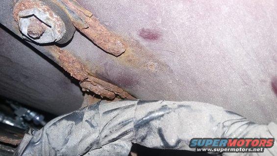Close-up of a rusted metal bracket attached to a vehicle's undercarriage, with a dirty wiring harness visible