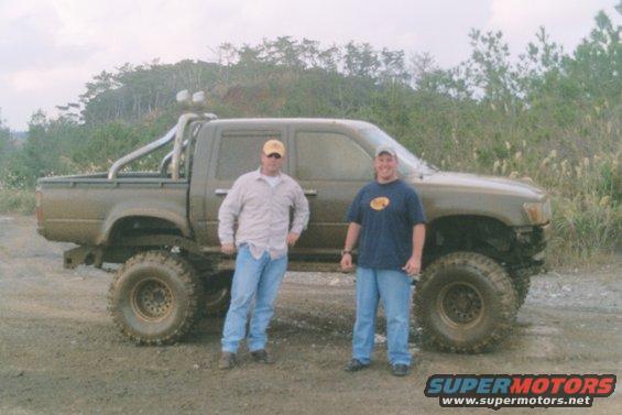Lifted pickup truck with roll bar and large tires, two men standing beside it