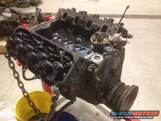 Engine block with valve covers removed, suspended by a chain over a yellow bucket