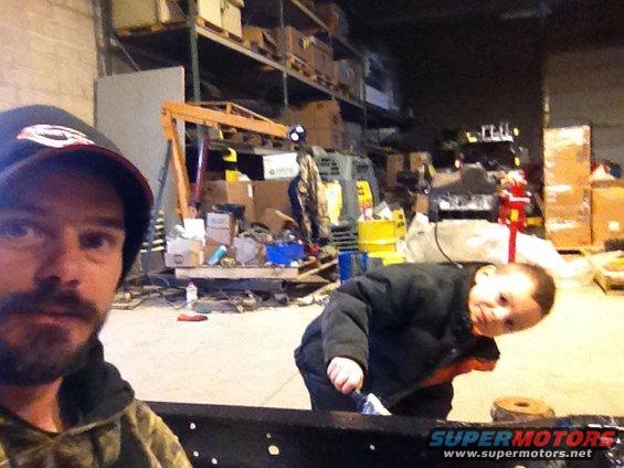 Man in a cap and camouflage jacket with a child in a workshop, shelves with boxes in background