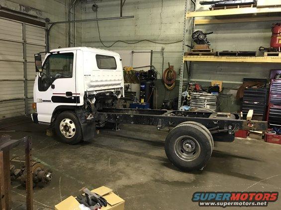 White truck chassis with dual rear wheels, viewed from the side in a workshop. The cab is intact, but the rear body is missing