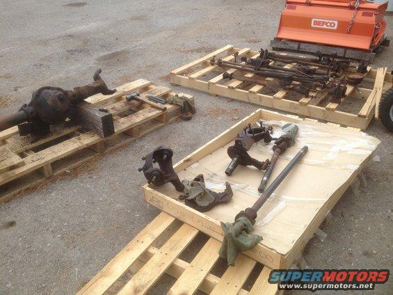 Vehicle axles and suspension parts on pallets, with an orange BEPCO attachment in background