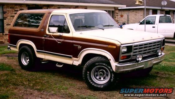 1980 Ford Bronco, two-tone brown and cream, parked on grass in front of a brick house. White pickup truck in background