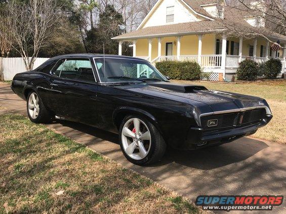 Black 1969 Mercury Cougar with a hood scoop and silver wheels parked in front of a yellow house