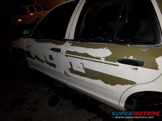 Side view of a white Ford Crown Victoria with peeling green and white decals. Another SUV is parked behind it