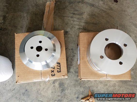 Two aluminum pulleys on cardboard boxes, one with four bolt holes and a central opening, the other with three grooves