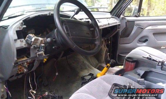 Vehicle interior with dashboard disassembled, wires exposed, and steering wheel visible. Gray seats and door panel are present