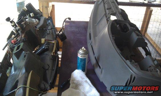 Two disassembled dashboard sections, one black and complex with wiring, the other gray and simpler, sit on a purple surface next to a can of spray paint and a white cloth