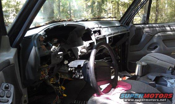 Interior of a vehicle with the dashboard removed, showing wires and components. Steering wheel and seats are visible