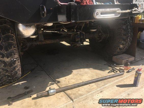 Rear axle and suspension of a lifted off-road vehicle, with a tie rod assembly removed and lying on the ground. Tools and a can of lubricant are nearby