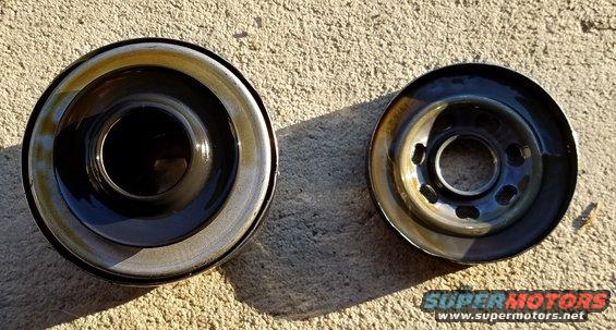 Two oil filters, one larger than the other, sit on a textured surface. The larger filter has a black center and a silver rim with oil residue. The smaller filter has a metal base with multiple bolt holes and oil residue