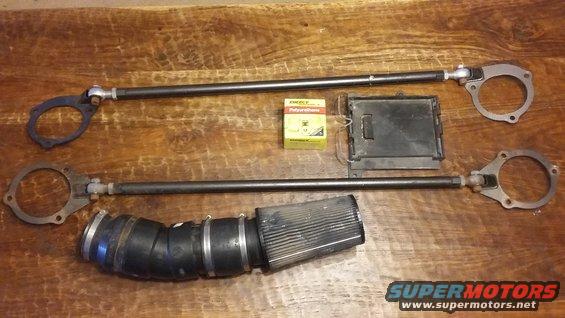 Two adjustable strut bars, an air intake assembly, a black electronic module, and a small yellow box of sealant on a wooden surface