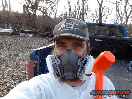 Man wearing a respirator and white protective suit, holding an orange mallet, standing in front of a blue pickup truck bed