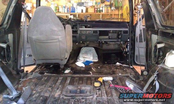 Interior of a disassembled vehicle, showing exposed floor, dashboard, and seats. Tools are scattered on the floor
