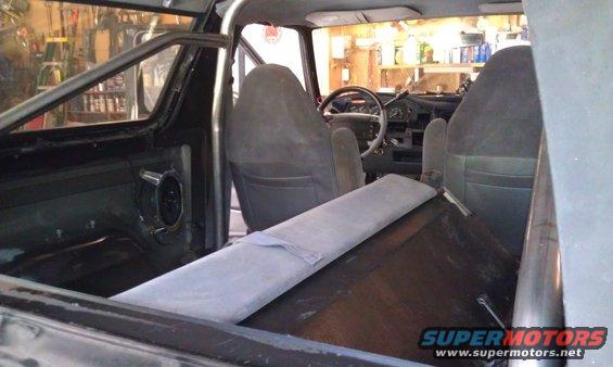 Interior view of a vehicle with bucket seats, a roll cage, and a dashboard. The rear cargo area has a padded cover
