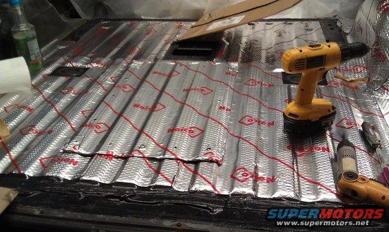 Car floor covered in silver insulation with red text, yellow drill, and tools