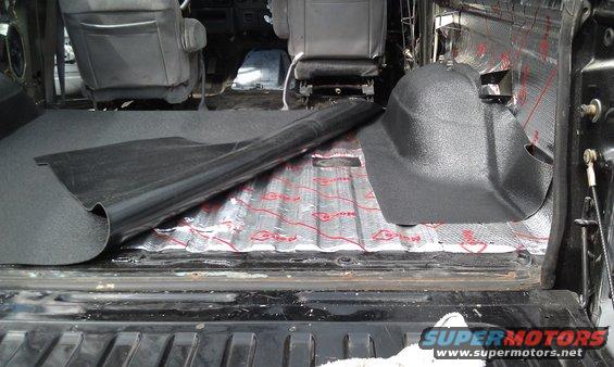 Vehicle interior with sound deadening material on the floor and insulation on the walls. A black plastic trim piece is visible