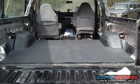 Interior of a vehicle showing new dark grey flooring installed in the cargo area and front seats visible. Another SUV is parked behind