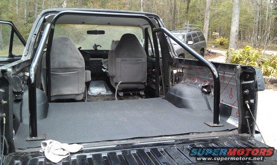 Black Ford Bronco interior with grey seats, roll cage, and carpeted bed. A white cloth is on the floor
