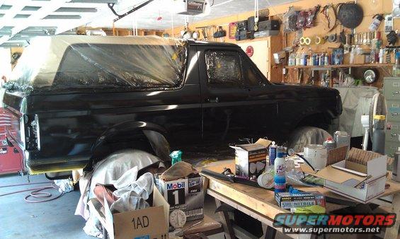 Black Ford Bronco in garage, covered in plastic sheeting, with paint supplies on a workbench