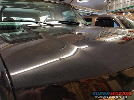 Dark SUV hood with a dent, windshield wipers, and an egg carton. Another SUV is visible behind it