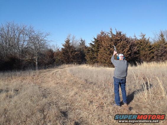 man shooting a rifle in a dry field with trees and blue sky
