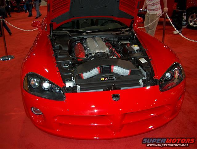 Red sports car, likely a Dodge Viper, with hood open showing the engine. Displayed indoors on a red carpet