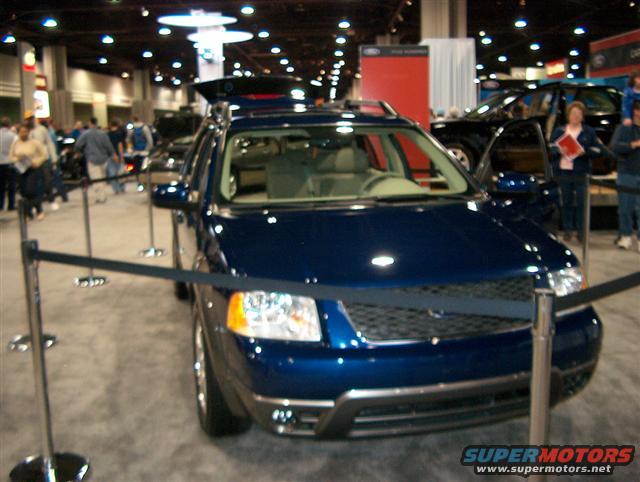 Blue SUV with its rear hatch open, displayed indoors at an auto show with other vehicles and people around
