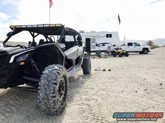 Side-by-side UTV parked in sand, with a trailer and pickup truck behind it