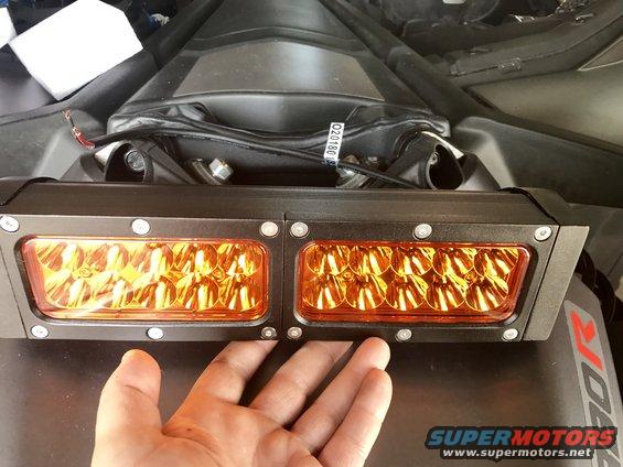 Hand holding a dual amber LED light bar mounted on a black surface with wiring visible