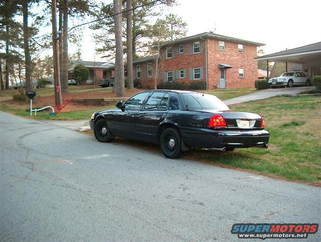 Black Ford Crown Victoria parked on a residential street. Another white car is visible under a carport