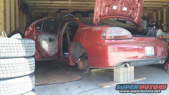 Red sedan with doors and trunk open, wheels removed, on blocks in a garage. License plate reads A89 7585